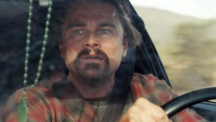 Leonardo DiCaprio looking concerned in the driver's seat of a car from One Battle After Another backtothepicture.net still